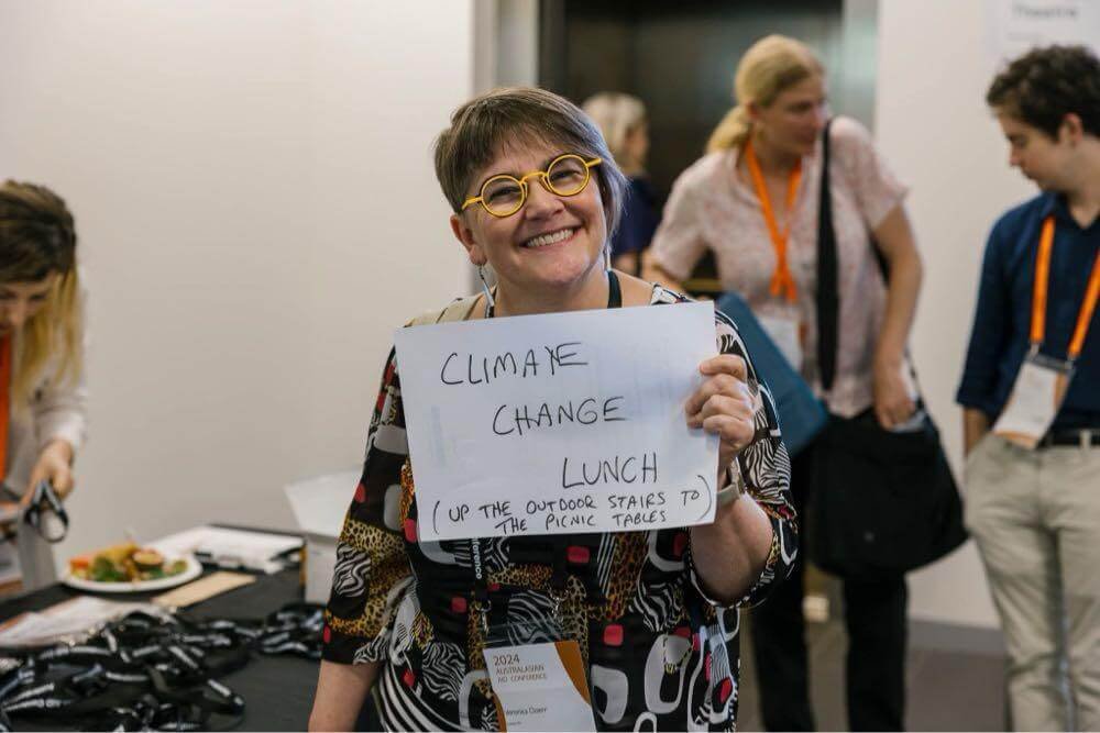 Veronica Doerr at the AAC, holding up a handwritten sign that says 'Climate Change Lunch! Up the outdoor stairs to the picnic tables'