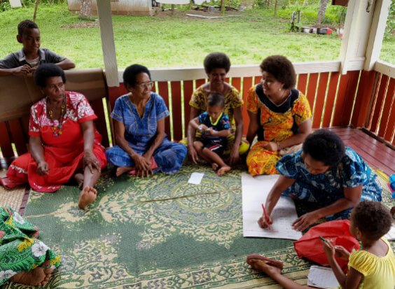 A group of Fijian women sitting in a circle on a veranda, partaking in a workshop, with one woman taking notes on butcher's paper
