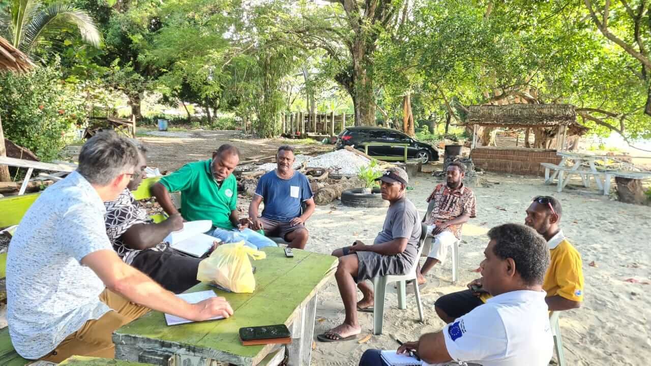 A focus group discussion with 7 participants around a table on sandy ground in Kaware, being facilitated by Dr Matt Allen