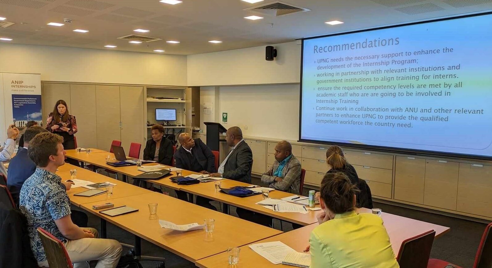 Sustineo CEO Tom Sloan seated at a large table with 8 other people visible around the table. Someone is presenting with a slide that says 'Recommendations'