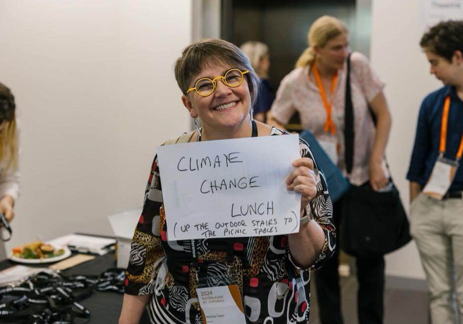 Veronica Doerr at the AAC, holding up a handwritten sign that says 'Climate Change Lunch! Up the outdoor stairs to the picnic tables'