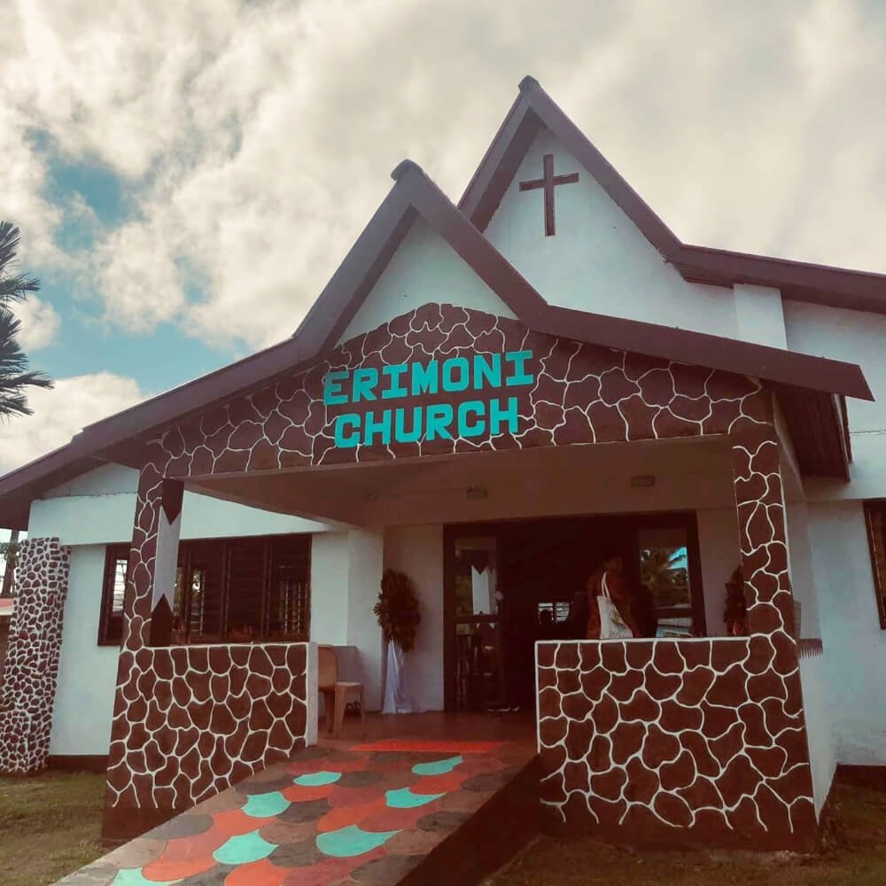Photo of 'Erimoni Church' with white walls, high pitched roof, colourful tiled path leading up to the entrance.