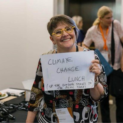 Veronica Doerr at the AAC, holding up a handwritten sign that says 'Climate Change Lunch! Up the outdoor stairs to the picnic tables'